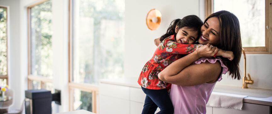 A mother laughs while holding her smiling daughter in the kitchen.