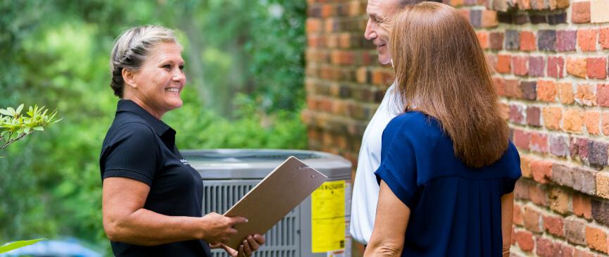 An American Standard HVAC dealer holds a clipboard as she explains HVAC troubleshooting steps to a couple.